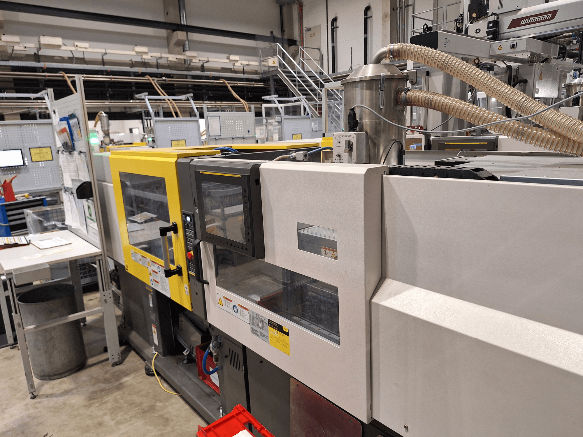 Vorderansicht einer mehrfarbigen Spritzgießmaschine mit gelben und grauen Teilen, mit Bedienfeld und Absauganlage.